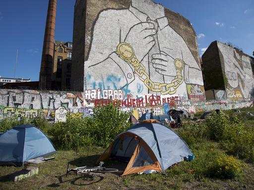 Zelte stehen auf einem kleinen Grünstreifen vor einer Häuserwand. Das Grafitti verweist auf die steigenden Mietpreise in Berlin.
