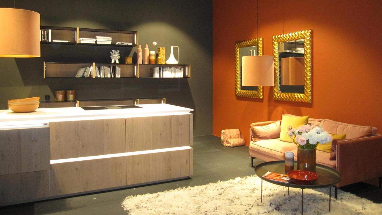 Zu sehen ist eine in warmen Farben gestaltete opulente Wohnküche mit Teppich, Samtsofa, großem Lampenschirm, Bücheregalen und Spiegeln in goldfarbenen Rahmen. Zu sehen ist eine in warmen Farben gestaltete opulente Wohnküche mit Teppich, Samtsofa, großem Lampenschirm, Bücheregalen und Spiegeln in goldfarbenen Rahmen.