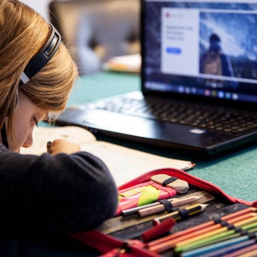 Ein Grundschulkind mit Headset sitzt in einem Wohnzimmer am Tisch und macht Homeschooling. Auf dem Tisch steht ein aufgeklappter Laptop. Das Kind löst die Hausaufgaben in einem Schulbuch. Auf dem Tisch liegen weitere Utensilien für die Schule.