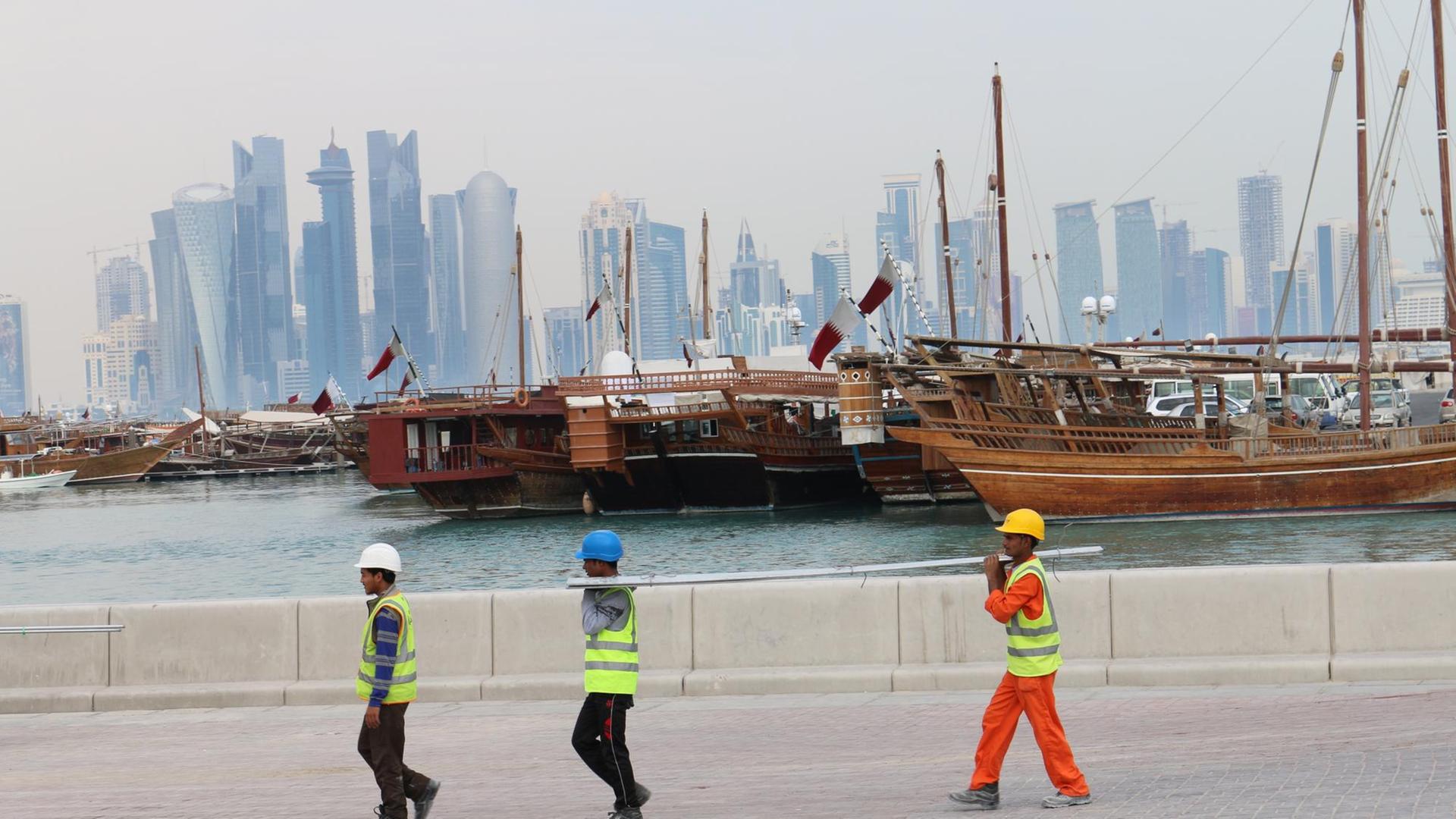 Drei Bauarbeiter gehen am Dau-Hafen entlang mit der Skyline der katarischen Hauptstadt Doha im Hintergrund. Drei Bauarbeiter gehen am Dau-Hafen entlang mit der Skyline der katarischen Hauptstadt Doha im Hintergrund.