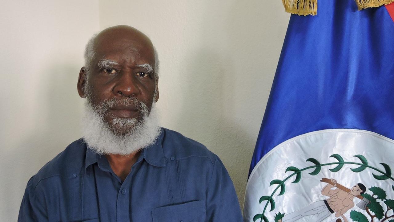 Ein Mann mit weißem Bart und blauem Hemd steht neben der Flagge von Belize. Ein Mann mit weißem Bart und blauem Hemd steht neben der Flagge von Belize.