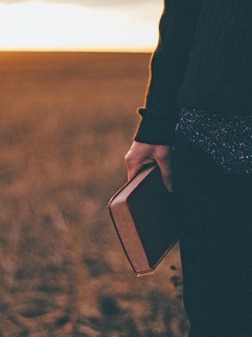 Jemand steht mit einem Buch in der Hand im Sonnenuntergang auf einem Feld.