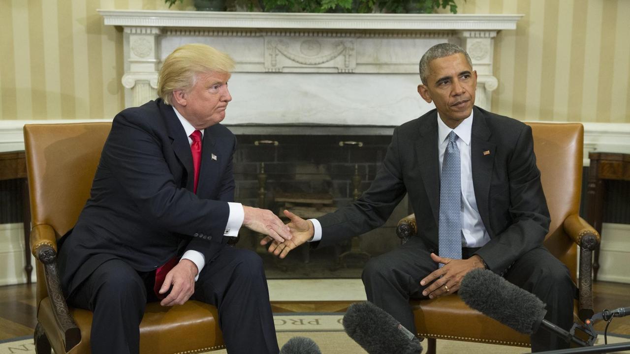 Donald Trump und Barack Obama reichen sich die Hände. Beide sitzen auf Stühlen im Oval Office, umringt von Mikrofonen.