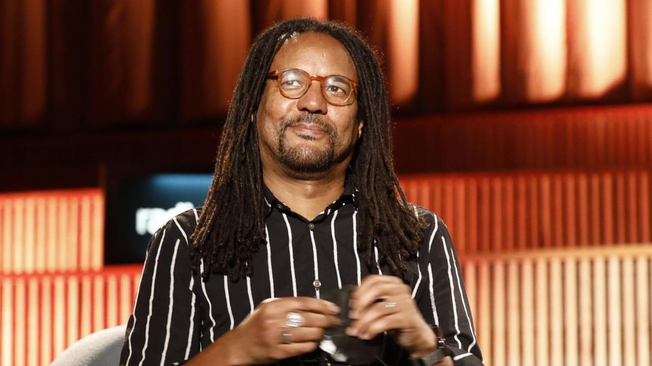 Der US-amerikanische Schriftsteller Colson Whitehead sitzt bei einer Lesung in einem schwarzweiß gestreiften Hemd in einem grauen Sessel. Der US-amerikanische Schriftsteller Colson Whitehead sitzt bei einer Lesung in einem schwarzweiß gestreiften Hemd in einem grauen Sessel.