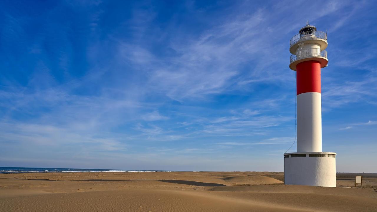 Ein rot-weißer Leuchturm steht am Strand von Deltebre, im Hintergrund ist das Meer zu sehen.