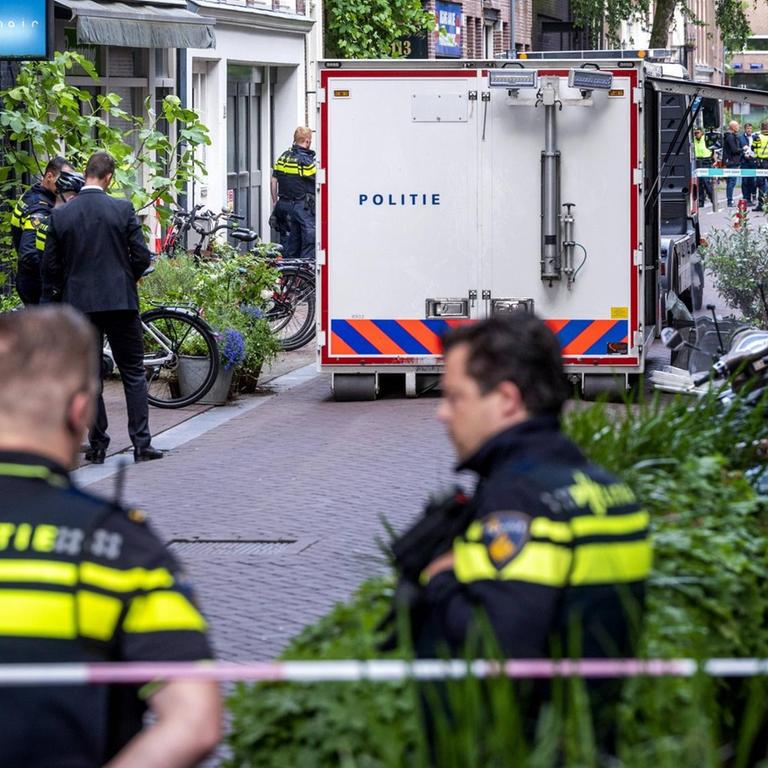 Zwei Polizisten stehen an einem Absperrband in einer Straße in Amsterdam. Im Hintergrund ist ein Polizeifahrzeug geparkt.