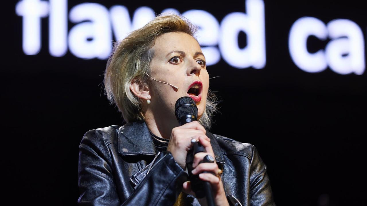 Eine Frau mit blonden Haaren und schwarzer Lederjacke spricht in ein Mikrophon. Hinter ihr ist ein großer Bildschirm auf einer Bühne zu sehen.