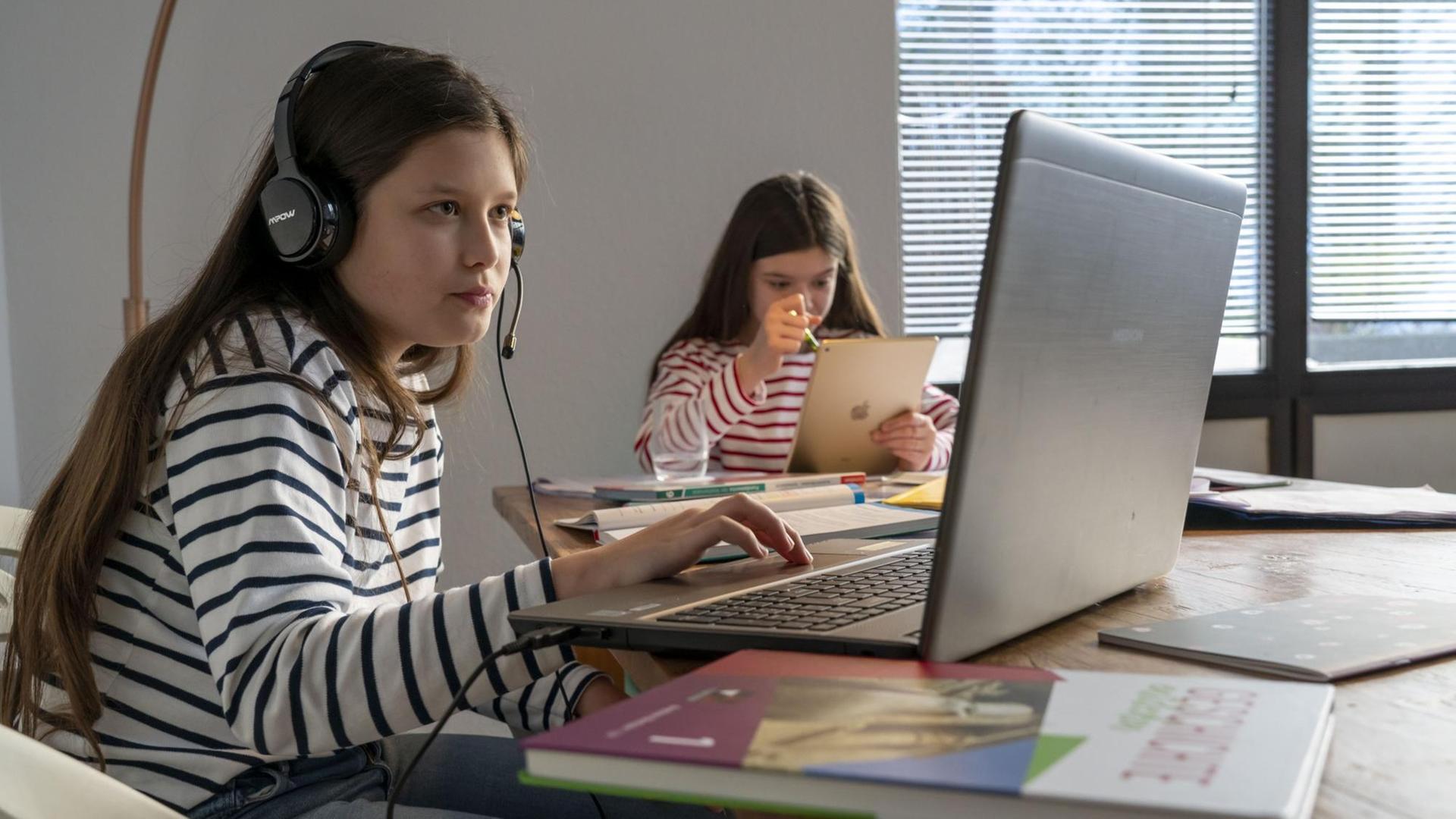 Homeschooling während des Lockdowns im Januar 2021: Ein Mädchen sitzt an einem Tisch vor einem Laptop im Videounterricht, neben sich ein Schulbuch. Im Hintergrund lernt ein weiteres Mädchen mit einem Tablet. Homeschooling während des Lockdowns im Januar 2021: Ein Mädchen sitzt an einem Tisch vor einem Laptop im Videounterricht, neben sich ein Schulbuch. Im Hintergrund lernt ein weiteres Mädchen mit einem Tablet.