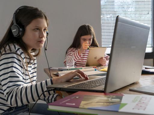 Homeschooling während des Lockdowns im Januar 2021: Ein Mädchen sitzt an einem Tisch vor einem Laptop im Videounterricht, neben sich ein Schulbuch. Im Hintergrund lernt ein weiteres Mädchen mit einem Tablet.