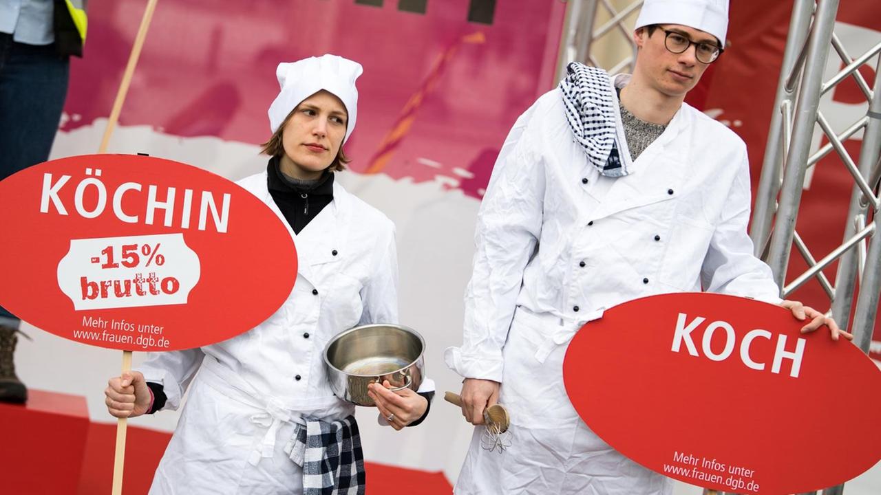 Demonstration für geschlechtergerechte Löhne: Eine Köchin und ein Koch zeigen mit Schildern, dass der Lohnunterschied zwischen beiden 15 Prozent beträgt.