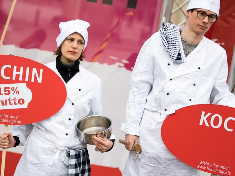 Demonstration für geschlechtergerechte Löhne: Eine Köchin und ein Koch zeigen mit Schildern, dass der Lohnunterschied zwischen beiden 15 Prozent beträgt.