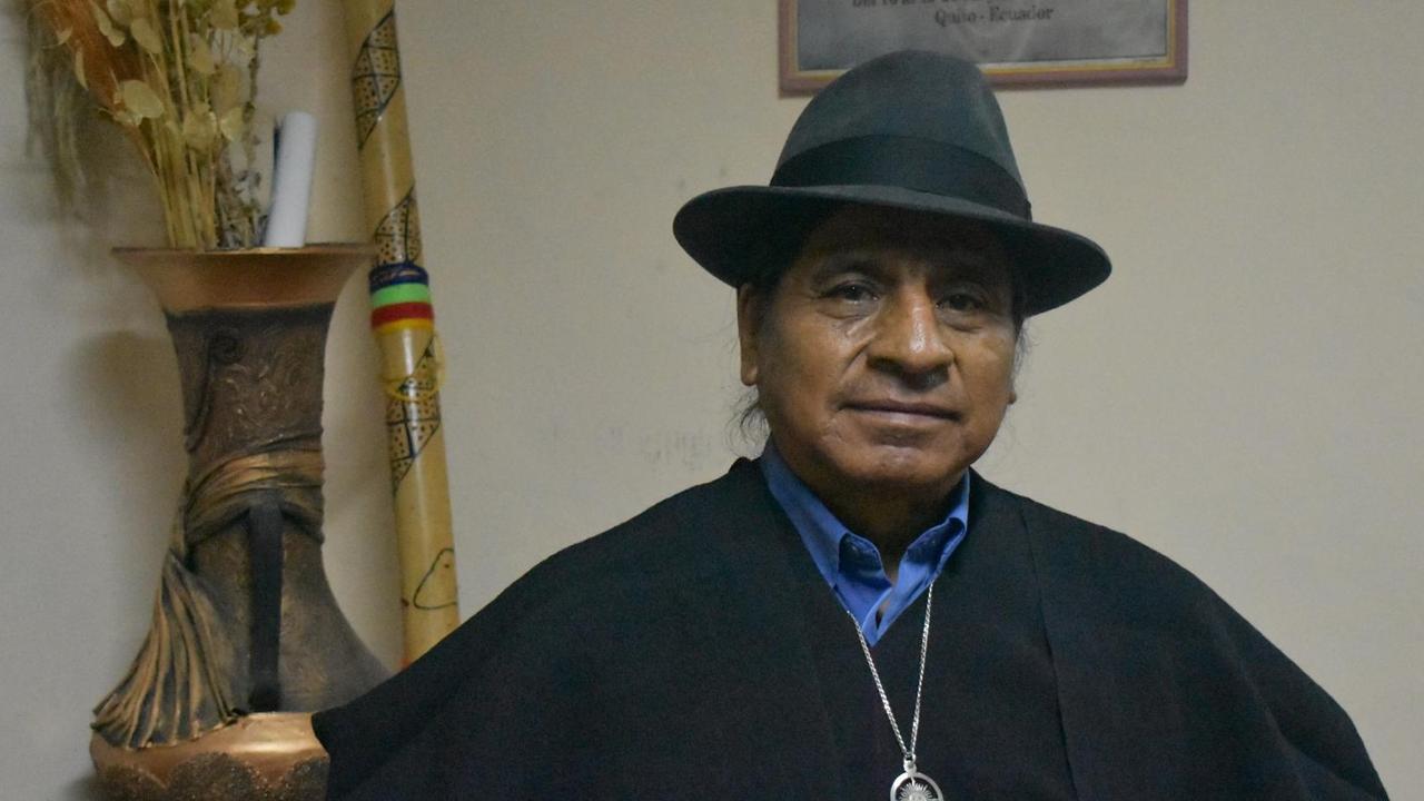 Luís Macas steht mit Poncho und Hut in einem Raum mit indigenen Accessoires. Luís Macas steht mit Poncho und Hut in einem Raum mit indigenen Accessoires.