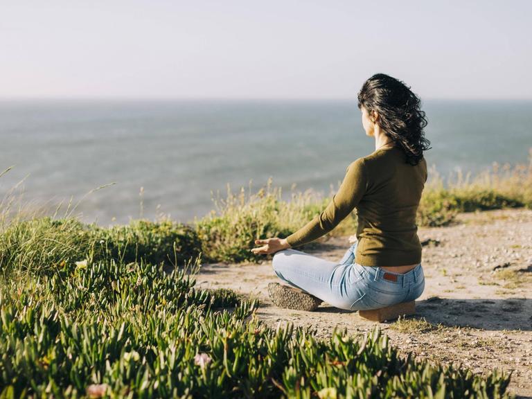 Eine Frau sitzt meditierend im Lotussitz auf dem Boden. Im Hintergrund sieht man das Meer.