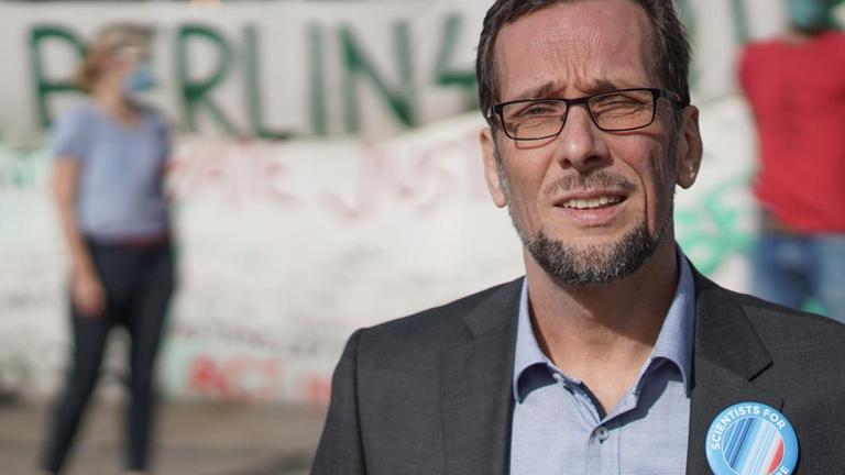 Ein Mann steht auf einem Platz, im Hintergrund sind Menschen und Transparente zu sehen. Der Mann im Vordergrund ist Volker Quaschning, Professor für regenerative Energiesysteme, er schaut in die Kamera. Ein Mann steht auf einem Platz, im Hintergrund sind Menschen und Transparente zu sehen. Der Mann im Vordergrund ist Volker Quaschning, Professor für regenerative Energiesysteme, er schaut in die Kamera.