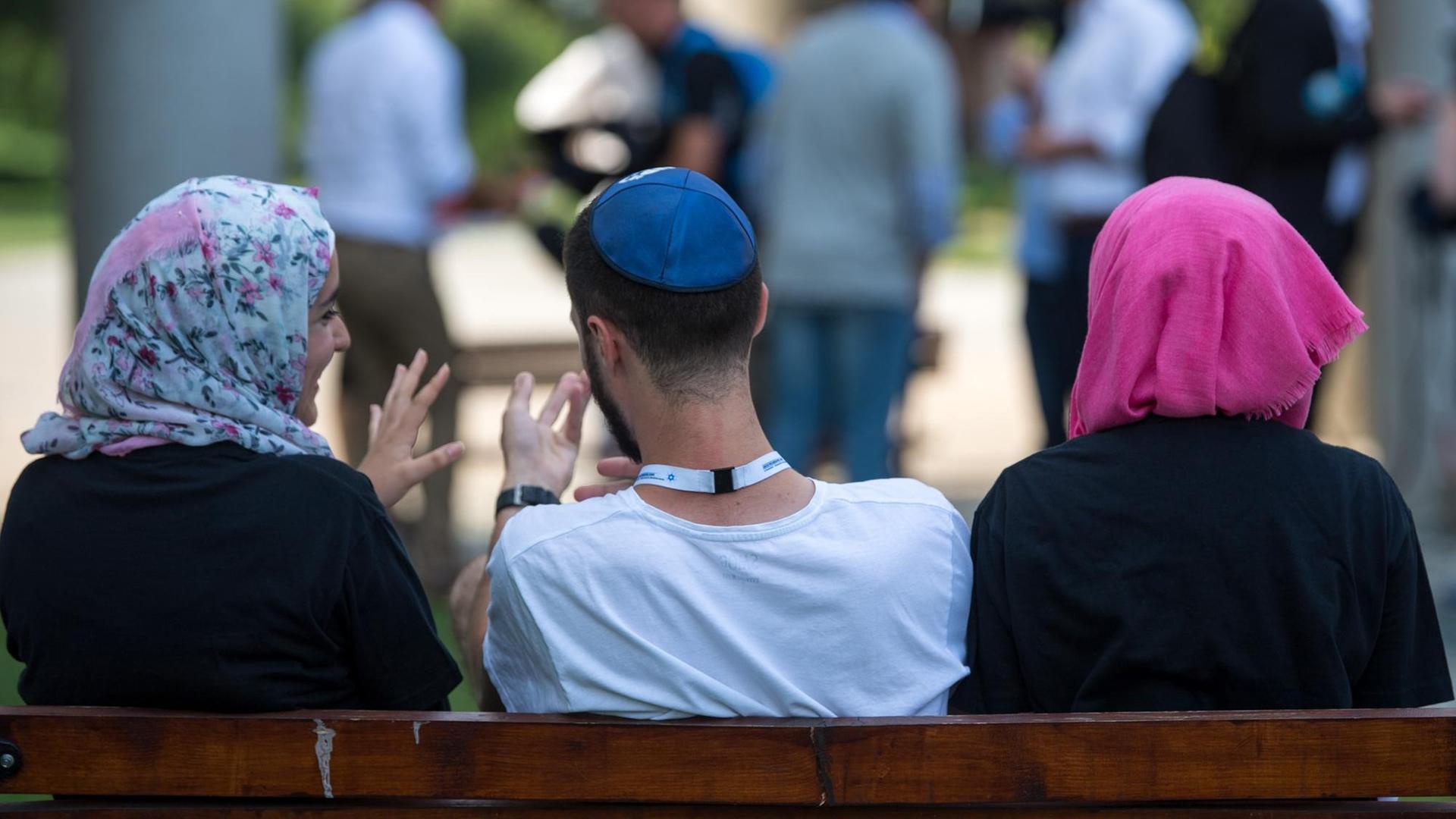 Symbolbild: Zwei junge Muslima mit Kopftuch und ein junger jüdischer Mann mit Kippa unterhalten sich miteinander. Sie befinden sich in der KZ-Gedenkstätte Auschwitz. Symbolbild: Zwei junge Muslima mit Kopftuch und ein junger jüdischer Mann mit Kippa unterhalten sich miteinander. Sie befinden sich in der KZ-Gedenkstätte Auschwitz.