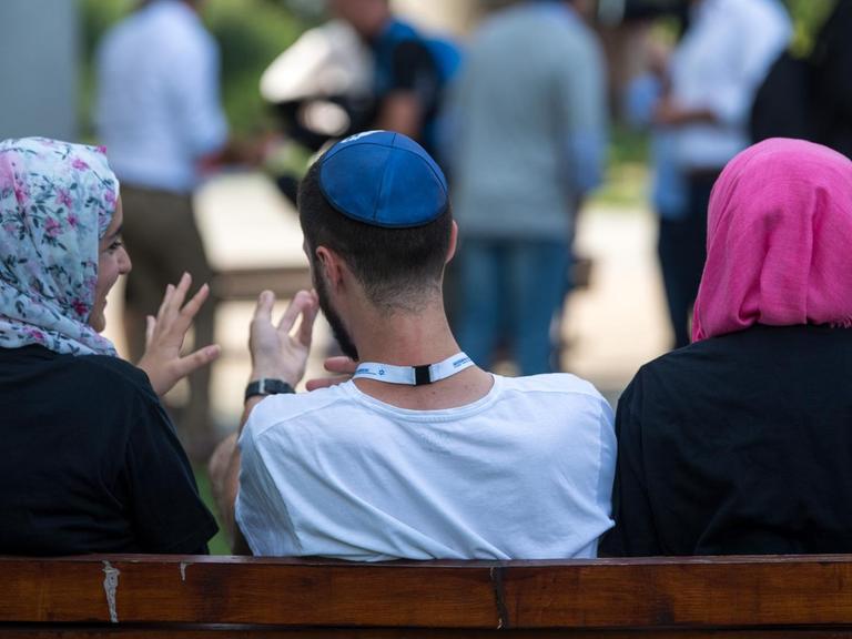 Symbolbild: Zwei junge Muslima mit Kopftuch und ein junger jüdischer Mann mit Kippa unterhalten sich miteinander. Sie befinden sich in der KZ-Gedenkstätte Auschwitz.