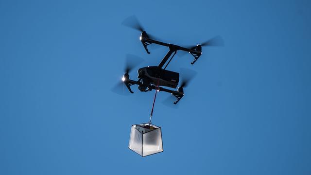 Eine Drohne fliegt mit einem kleinen Paket vor blauem Himmel.