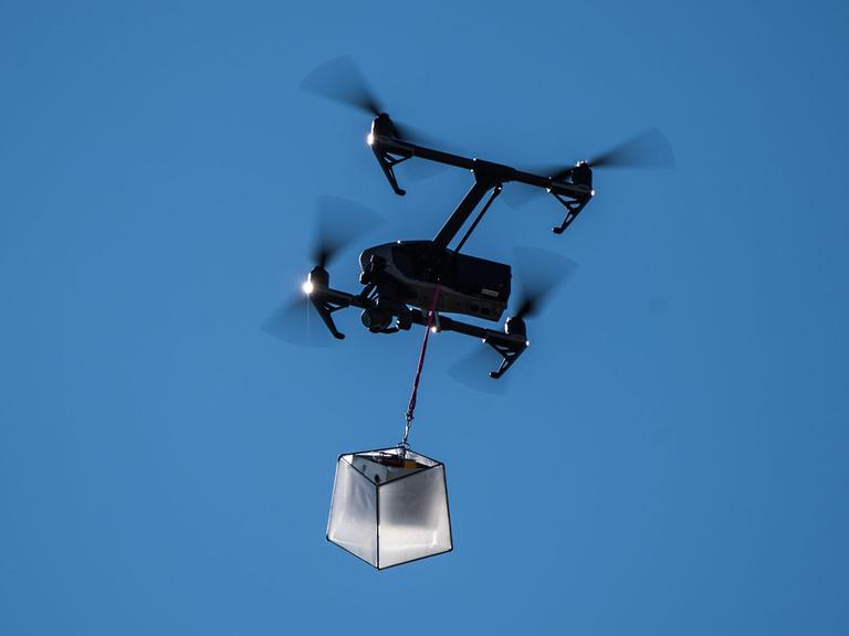 Eine Drohne fliegt mit einem kleinen Paket vor blauem Himmel.