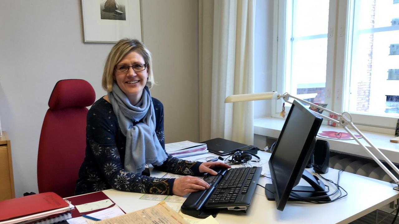 Sirkku Mietinen, Leiterin der Altenpflege der Kommune Tampere sitzt mit Pulli und Schal an ihrem Schreibtisch auf einem dunkelroten Stuhl, die Hände auf der Tastatur und lächelt in die Kamera.