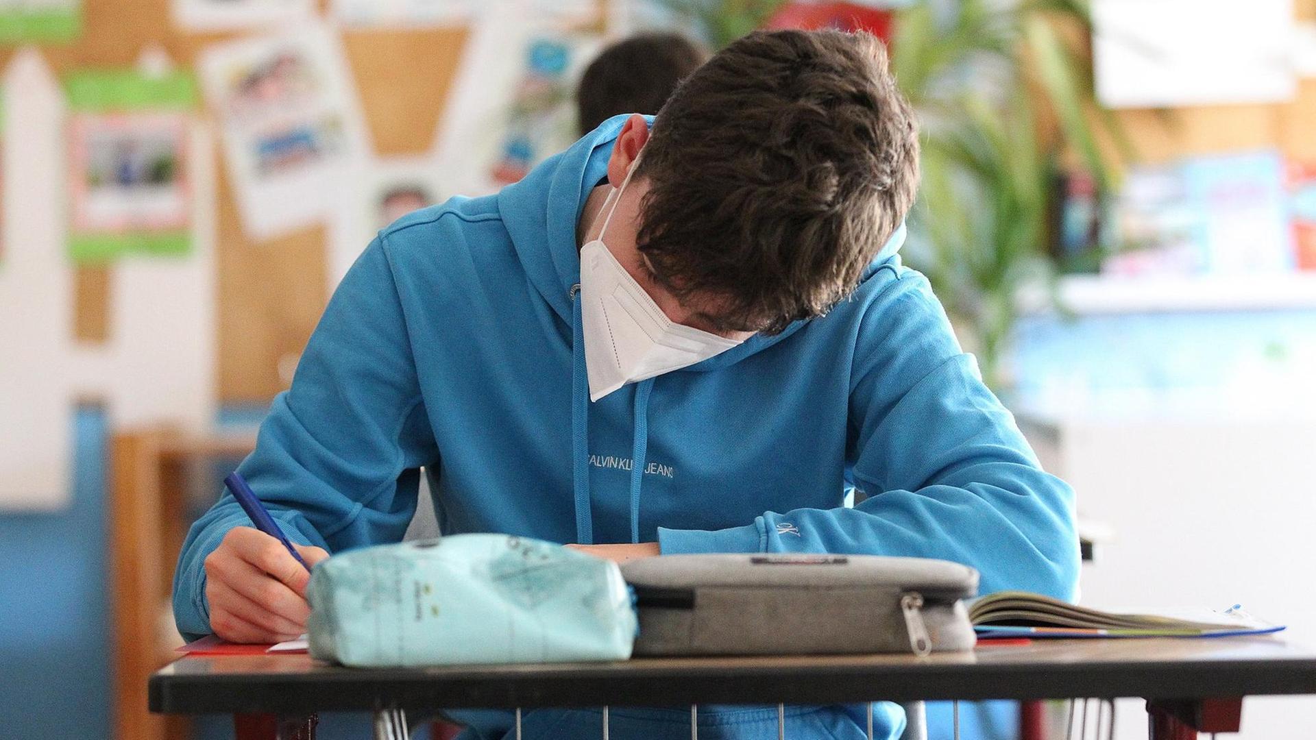 Ein Schüler sitzt mit einem Mundschutz im Klassenzimmer und schreibt. Ein Schüler sitzt mit einem Mundschutz im Klassenzimmer und schreibt.