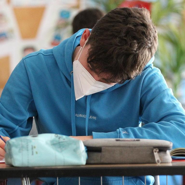 Ein Schüler sitzt mit einem Mundschutz im Klassenzimmer und schreibt. 