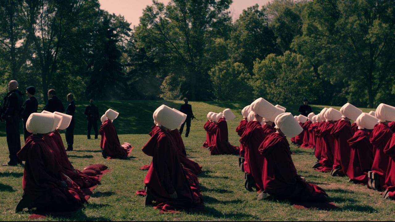 Szene aus "The Handmaid's Tale". Frauen in roten Roben und weißen Häubchen auf grüner Wiese. Dahinter Wachmänner. Szene aus "The Handmaid's Tale". Frauen in roten Roben und weißen Häubchen auf grüner Wiese. Dahinter Wachmänner.