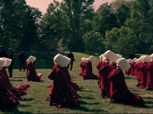 Szene aus "The Handmaid's Tale". Frauen in roten Roben und weißen Häubchen auf grüner Wiese. Dahinter Wachmänner.