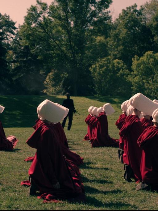 Szene aus "The Handmaid's Tale". Frauen in roten Roben und weißen Häubchen auf grüner Wiese. Dahinter Wachmänner.
