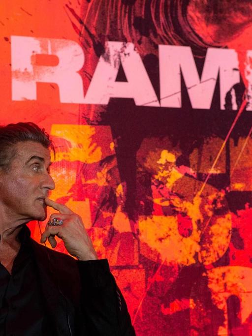 Der Schauspieler Sylvester Stallone sitzt in einem schwarzen Anzug vor einer Wand auf der "Rambo" geschrieben steht.