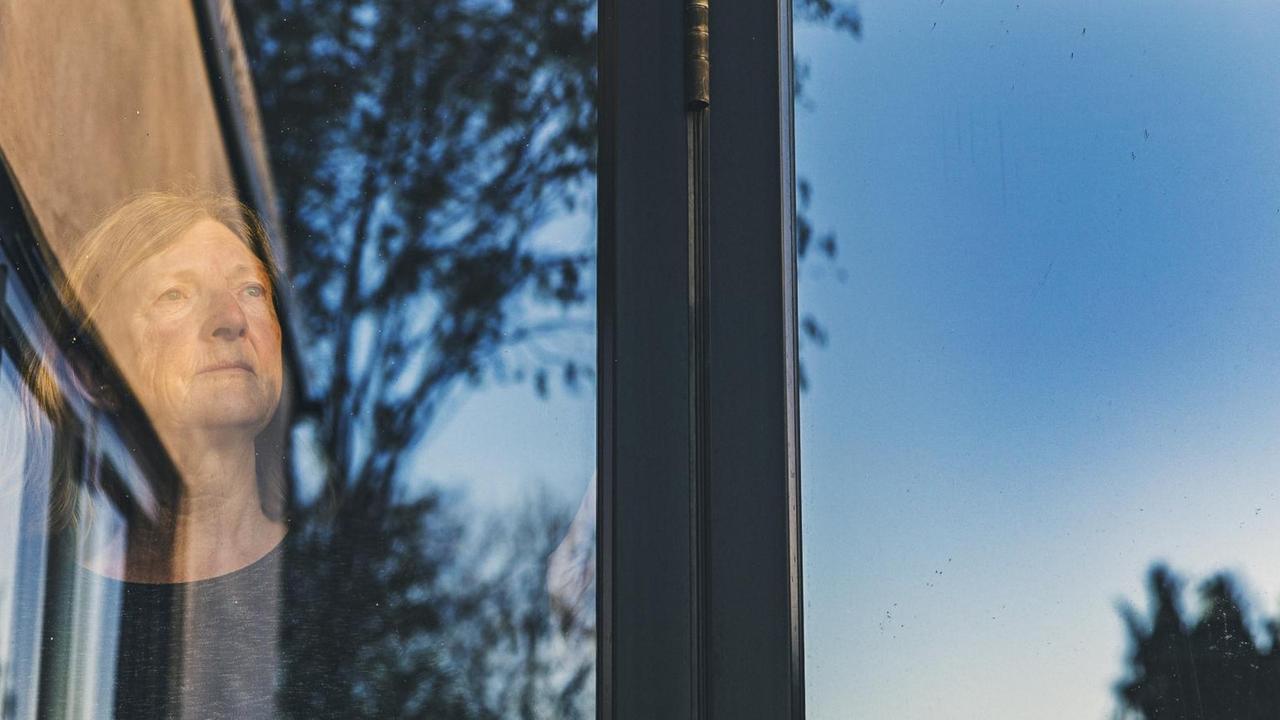 Eine ältere Frau steht am Fenster und blickt in die Ferne, in der Fenterscheibe spiegelnsich Himmel und Bäume. Eine ältere Frau steht am Fenster und blickt in die Ferne, in der Fenterscheibe spiegelnsich Himmel und Bäume.