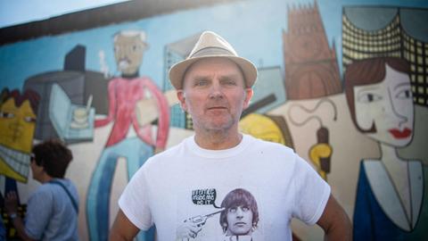 Der Künstler Jim Avignon steht vor seinem Werk an der East Side Gallery. Im Hintergrund sein Kunstwerk auf dem längsten noch erhaltenen Teilstück der Berliner Mauer.
