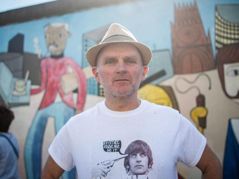 Der Künstler Jim Avignon steht vor seinem Werk an der East Side Gallery. Im Hintergrund sein Kunstwerk auf dem längsten noch erhaltenen Teilstück der Berliner Mauer.