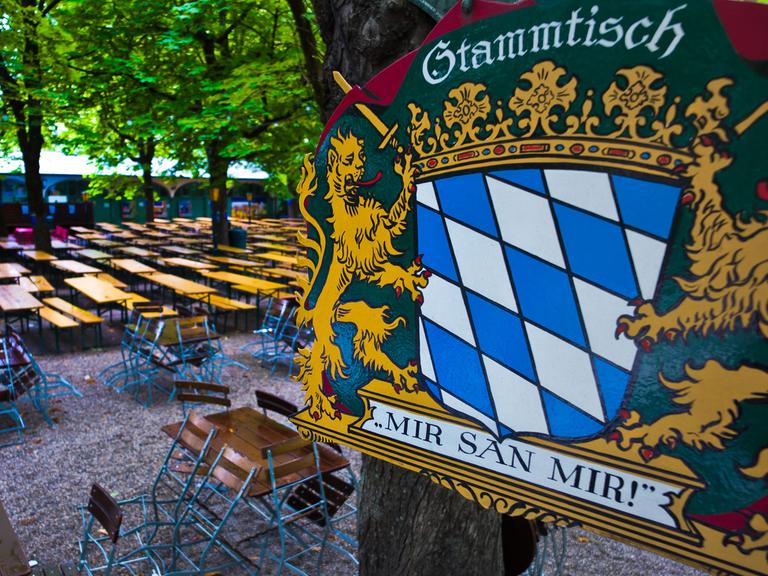 Ein Metallschild mit der Aufschrift "Stammtisch - Mir san mir!" hängt bei Regen in München über einem leeren Biergarten.