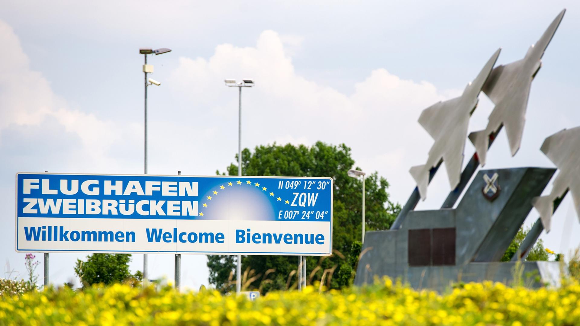 Schild mit der Aufschrift "Flughafen Zweibrücken. Willkommen, Welcome, Bienvenue" hinter einer Wiese mit gelben Blumen, daneben eine Skulptur aus drei metallenen Flugzeugen. Schild mit der Aufschrift "Flughafen Zweibrücken. Willkommen, Welcome, Bienvenue" hinter einer Wiese mit gelben Blumen, daneben eine Skulptur aus drei metallenen Flugzeugen.