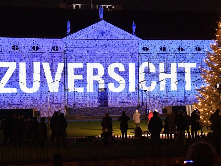 Im Rahmen der Lichtinstallation "Lichtblicke" ist auf das Schloss Bellevue in Berlin in Blau der Schriftzug "Zuversicht" projiziert. Davor steht bei einem Pressetermin unter anderen Bundespräsident Frank-Walter Steinmeier.