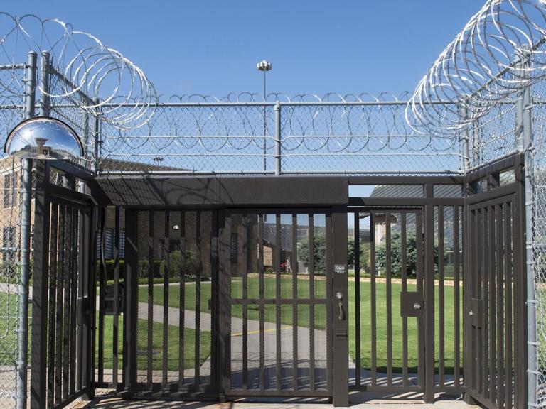 Blick auf den vergitterten Eingang eines Gefängnisses, die Zäune sind mit gerolltem Stacheldraht gesichert