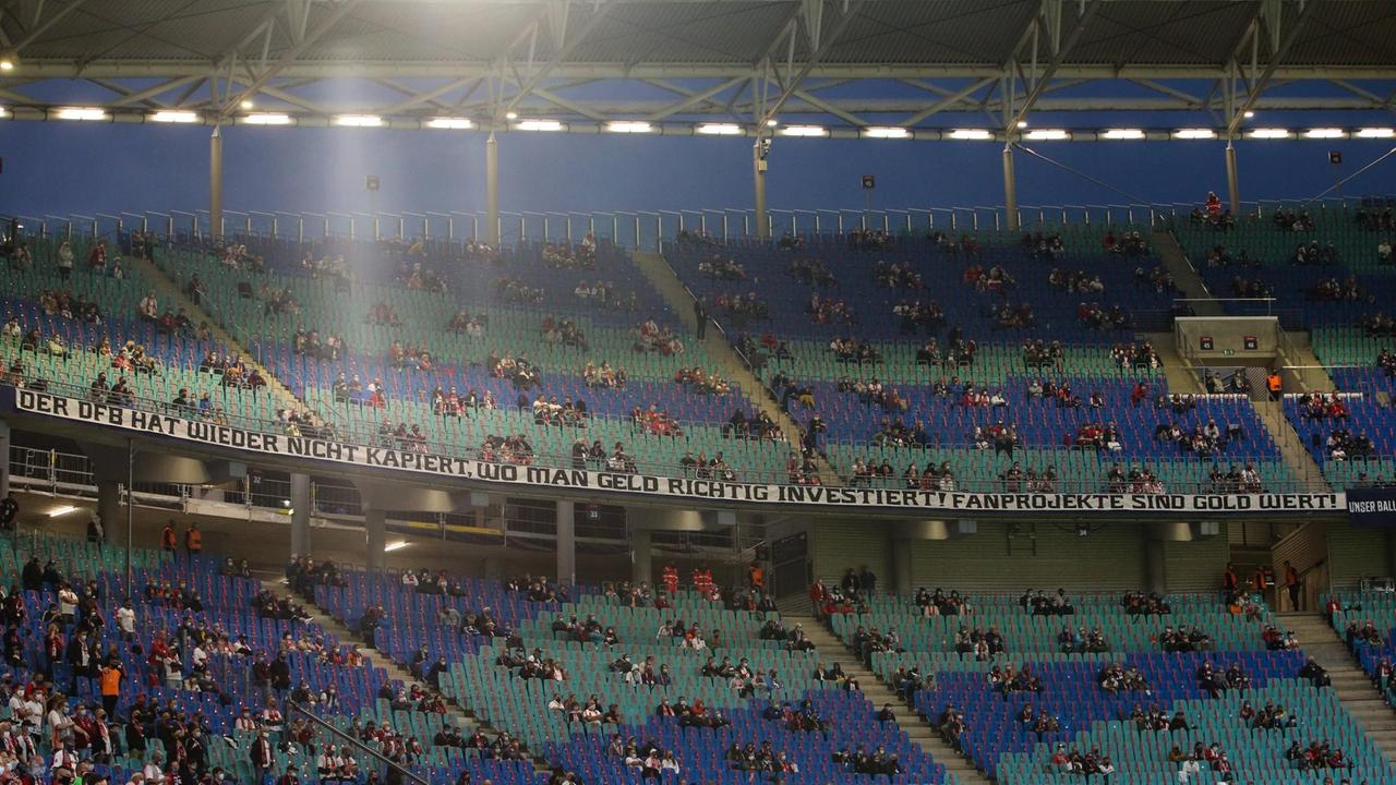 An einer Tribüne in einem Fußballstadion hängt ein Spruchband mit den Worten: "Der DFB hat wieder nicht kapiert, wo man Geld richtig investiert. Fanprojekte sind Gold wert!"
