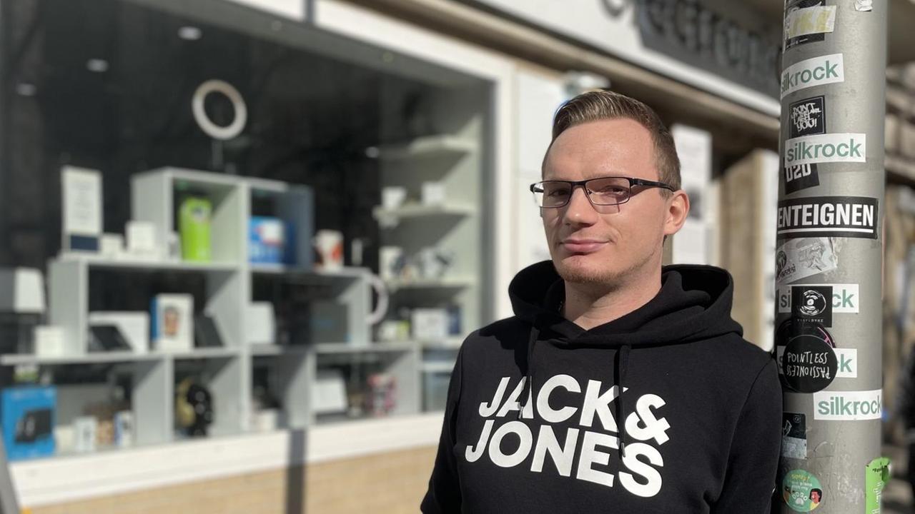 Ein junger Mann mit blondem Bürstenhaarschnitt und Hornbrille lehnt im schwarzen Kapuzenpulli an einer Straßenlaterne. Im Hintergrund das Schaufenster eines Computer- und Handyladens. Ein junger Mann mit blondem Bürstenhaarschnitt und Hornbrille lehnt im schwarzen Kapuzenpulli an einer Straßenlaterne. Im Hintergrund das Schaufenster eines Computer- und Handyladens.