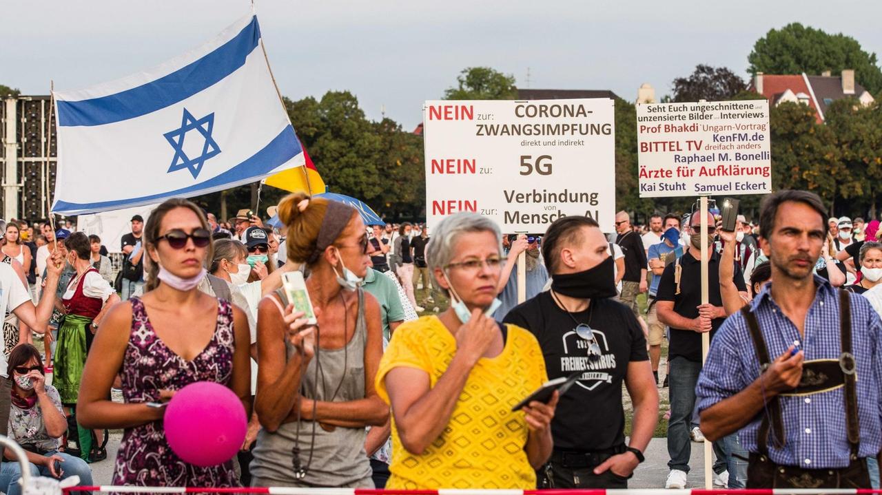 Demonstrierende, zum größten Teil ohne Mund-Nasen-Schutz, teilweise in Trachten stehen hinter einer Absperrung und tragen Schilder auf denen steht: Nein zur Corona-Zwangsimpung, Nein zu 5G, ... oder 'Ärzte für Aufklärung.' 
Auch eine israelische und eine deutsche Flagge sind zu sehen.
Ein Mann trägt ein Shirt mit der Aufschrift: 2020 Freiheitsvirus