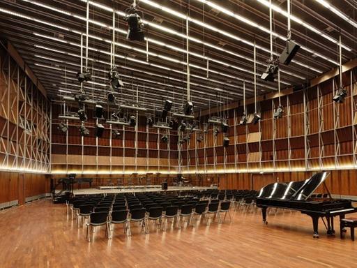 Zu sehen ist der menschenleere Kammermusiksaal im Deutschlandfunk-Gebäude. Ein großer Raum mit Parkett ausgelegt, darauf Stuhlreihen und davor ein Konzertpiano. Von der Decke hängen schwarze Scheinwerfer.