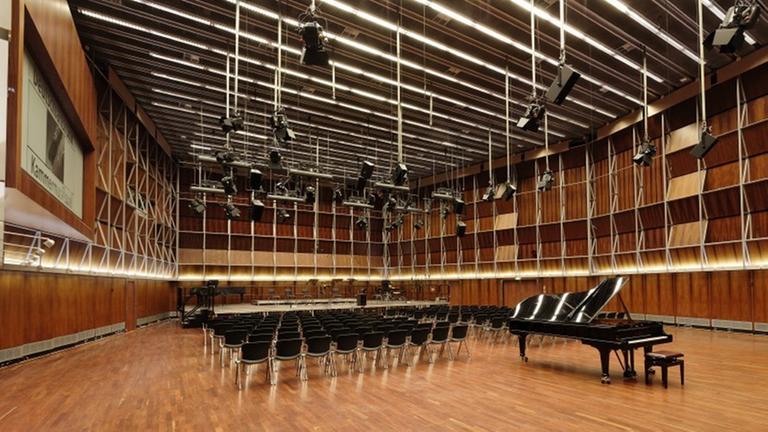 Zu sehen ist der menschenleere Kammermusiksaal im Deutschlandfunk-Gebäude. Ein großer Raum mit Parkett ausgelegt, darauf Stuhlreihen und davor ein Konzertpiano. Von der Decke hängen schwarze Scheinwerfer. Zu sehen ist der menschenleere Kammermusiksaal im Deutschlandfunk-Gebäude. Ein großer Raum mit Parkett ausgelegt, darauf Stuhlreihen und davor ein Konzertpiano. Von der Decke hängen schwarze Scheinwerfer.