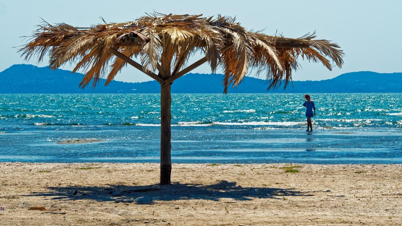 Im Vordergrund ein Sandstrand mit einem Sonnenschirm, dahinter türkis glitzerndes Meer, im flachen Wasser läuft eine Person.