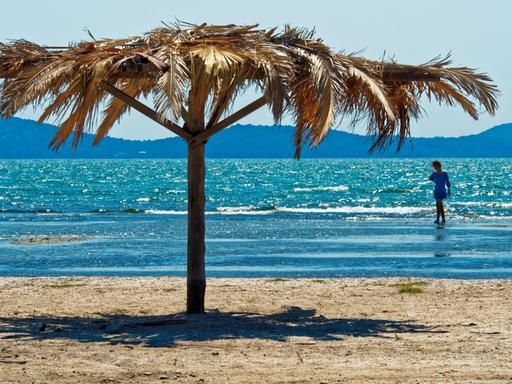 Im Vordergrund ein Sandstrand mit einem Sonnenschirm, dahinter türkis glitzerndes Meer, im flachen Wasser läuft eine Person.