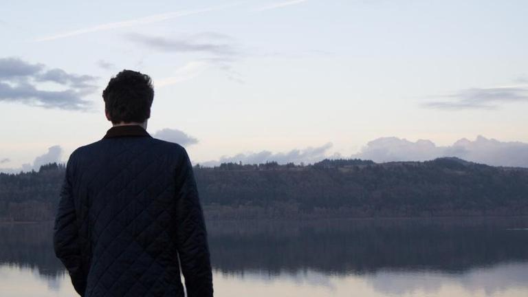 Ein Mann steht mit dem Rücken zum Betrachter vor einem See. Am Himmel sind Kondenstreifen zu sehen.

