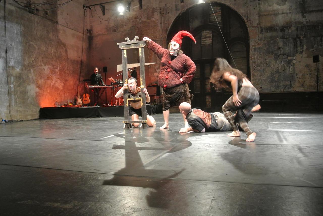 Vier Schauspieler sind auf der Bühne- Eine Person hält ihren Kopf in einer Guillotine, welche von einer weiteren, maskierten Person mit roter Zipfelmütze bedient wird. Am Boden liegt ein Mann und schaut gespannt dem Spektakel zu. Eine Frau scheint in die Richtung des Geschehens zu laufen, sie ist nur von hinten zu sehen. Vier Schauspieler sind auf der Bühne- Eine Person hält ihren Kopf in einer Guillotine, welche von einer weiteren, maskierten Person mit roter Zipfelmütze bedient wird. Am Boden liegt ein Mann und schaut gespannt dem Spektakel zu. Eine Frau scheint in die Richtung des Geschehens zu laufen, sie ist nur von hinten zu sehen.