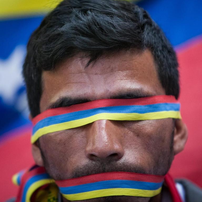 Ein Mann trägt Bänder in den Landesfarben Venezuelas über den Augen und über dem Mund. Im Hintergrund die Flagge des Landes.