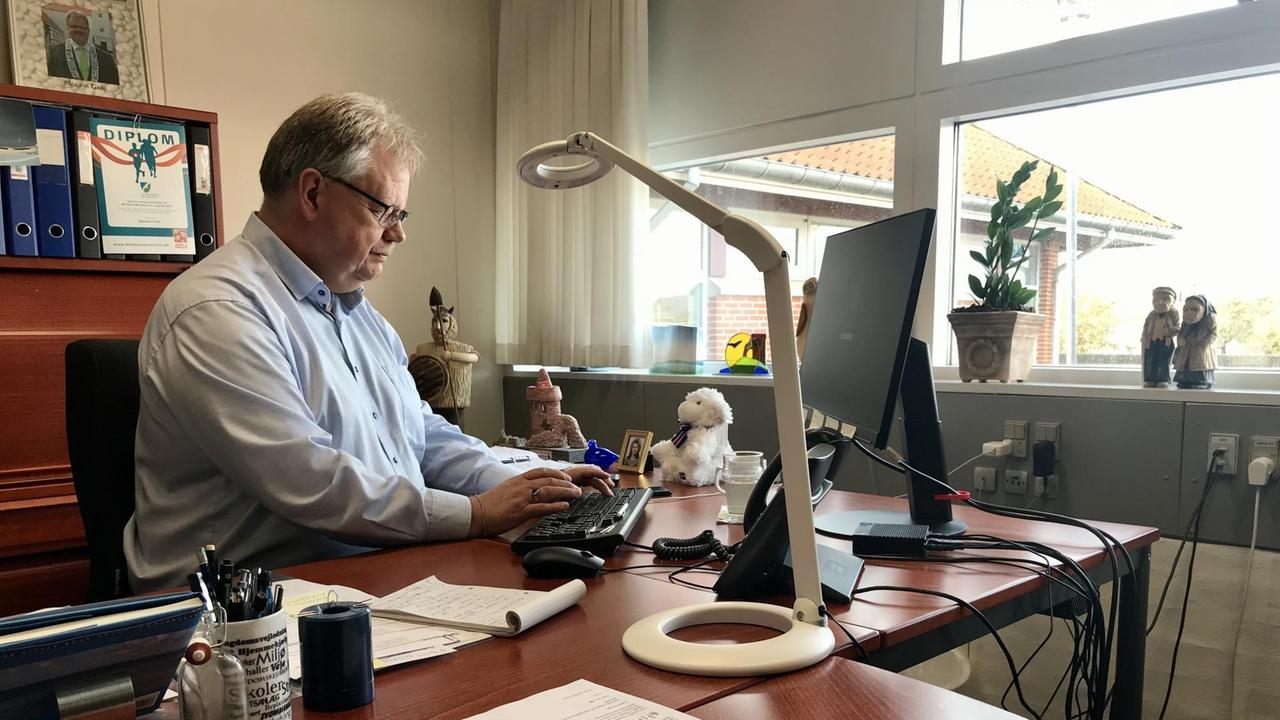 Ein weißhaariger Mann mit Brille tippt in seinem Büro mit Fensterblick am Schreibtisch sitzend auf seiner PC-Tastatur.
