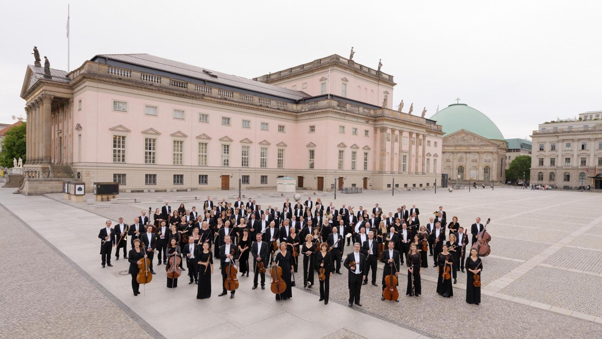 Die Musiker stehen mit ihren Instrumenten auf dem großen Bebelplatz, im Hintergrund ihr restauriertes Opernhaus Unter den Linden in Berlin.