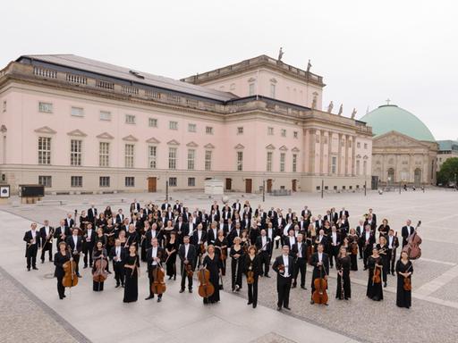 Die Musiker stehen mit ihren Instrumenten auf dem großen Bebelplatz, im Hintergrund ihr restauriertes Opernhaus Unter den Linden in Berlin.