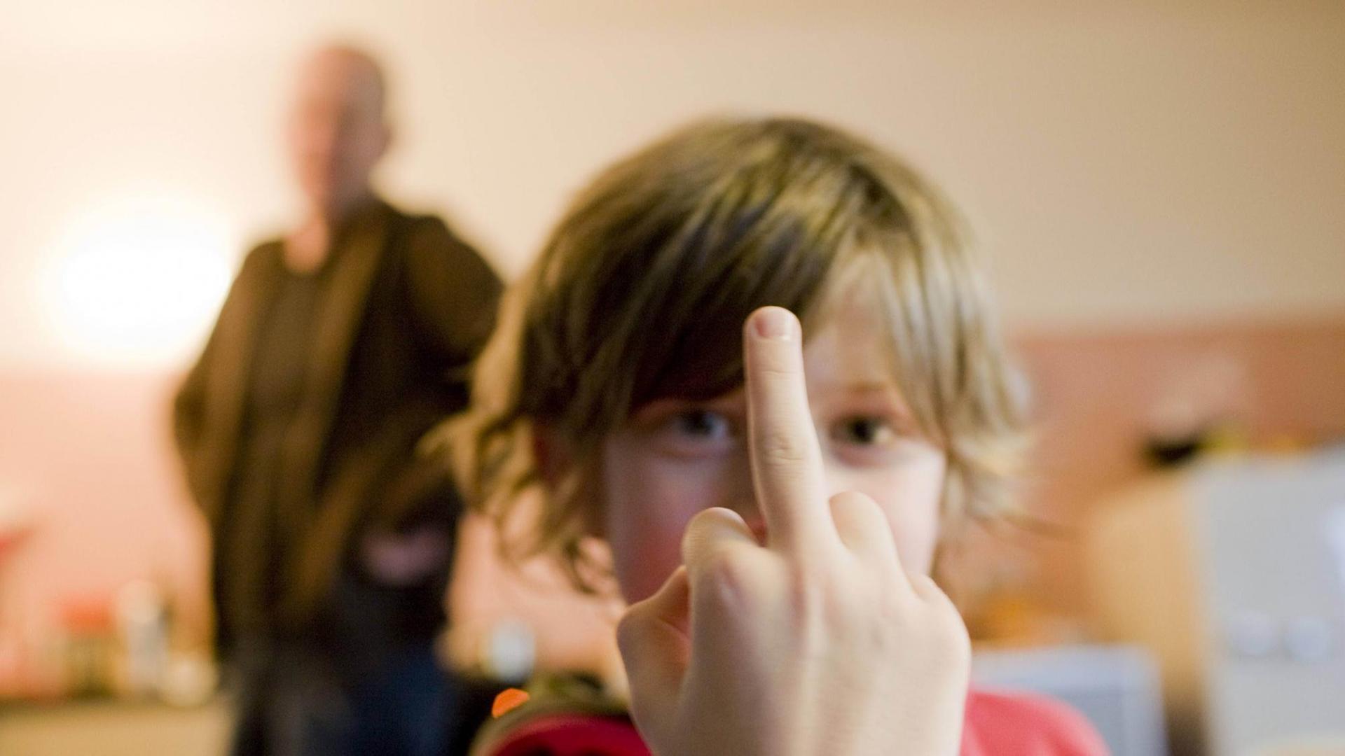 Ein Junge zeigt den Stinkefinger, im Hintergrund ist verschwommen ein erwachsener Mann zu sehen. Ein Junge zeigt den Stinkefinger, im Hintergrund ist verschwommen ein erwachsener Mann zu sehen.